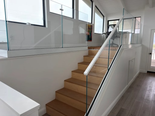 Frameless glass guards line an oak stair and the loft above under a white vaulted ceiling with black-frame windows.