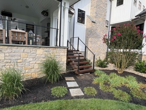 Black cable railing lines hardwood exterior stairs rising from a landscaped bed to a stone-based covered porch.