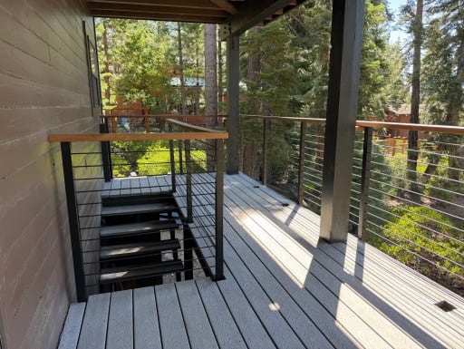 A covered gray deck with wood-capped cable railing and an open metal stair sits among pines.