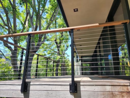 Black fascia-mounted posts with wood cap and cables line an elevated deck beneath a modern roof overhang.