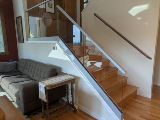 Frameless glass railing with a slim top cap follows a wood stair above a living room sofa.