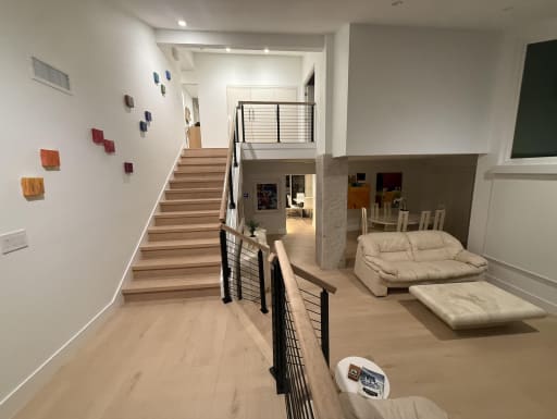 Black cable railing with wood handrails borders a light oak stair rising from a bright living area.