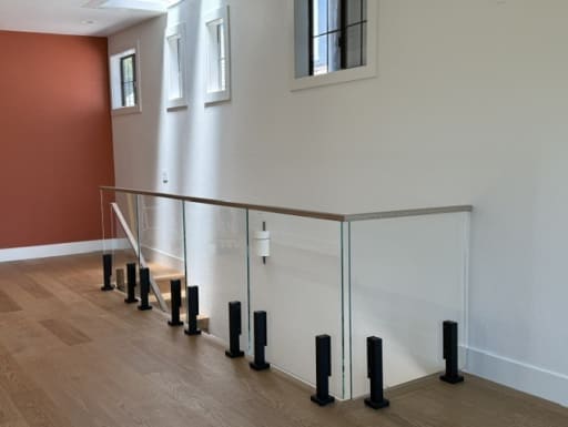 Black talons hold frameless glass with a white oak cap around a stair opening in a bright upstairs hall.