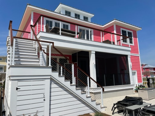 Bright pink coastal home with a white exterior stair and balconies trimmed in cable railing and a wood top rail.
