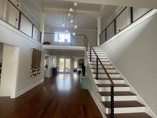 Black cable railing with walnut handrail on a white stair leading to a balcony in a long entry hall.