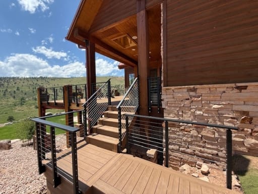 Black cable railing follows exterior deck stairs and landing beside a stone and wood mountain home.