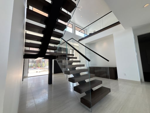 Underside view of dark wood floating treads on black mono stringers with frameless glass railing in a bright white hall.