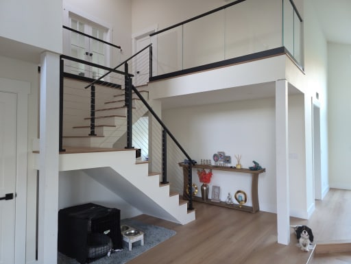 Black cable stair railing leads to a glass-on-base-rail loft in a bright white foyer; a small dog sits by the hall.
