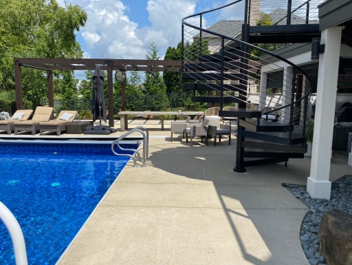 Black steel spiral stair with curved cable railing rises from a pool deck to a second-story balcony.