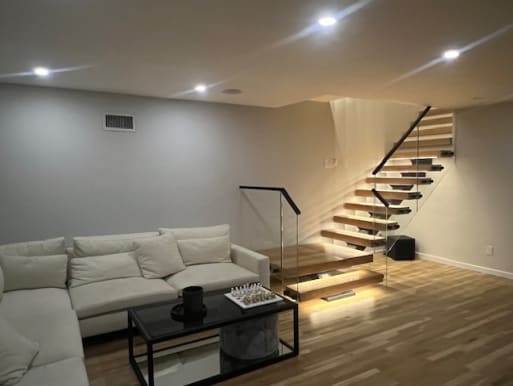 Floating oak treads with under-tread LEDs and glass railing rise from a cozy family room.