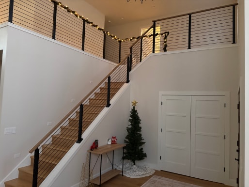 Black posts with cable infill and a wood cap rail rise along a white stair wall to a loft balcony.
