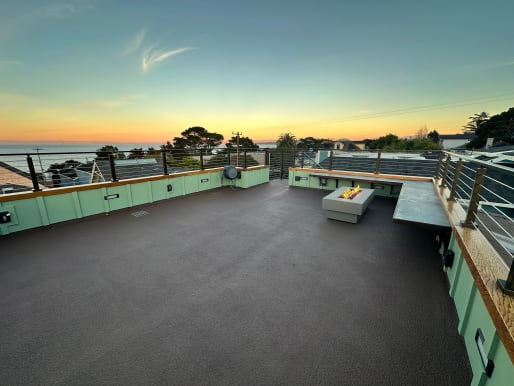 Cable railing lines a rooftop deck with a fire table and built-in bar under a sunset sky with ocean views.