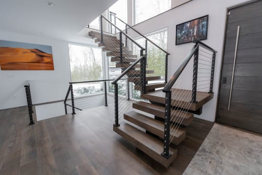 Stained oak floating treads with black cable railing turn at a landing beside tall windows.