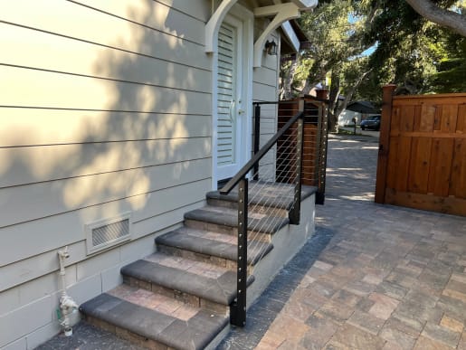 Black-post cable railing follows stone entry steps beside a white door and a cedar gate on a paver courtyard.