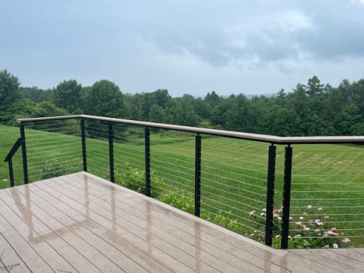 Black side-mount cable railing with a wide composite beverage rail edges a deck overlooking a green meadow.