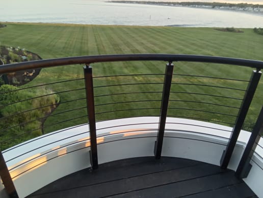 A curved round metal handrail tops black cable railing on a rounded balcony overlooking a lawn and the sea.