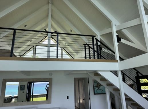 Black rod railing lines a white-trimmed loft and stair beneath an exposed-beam vaulted ceiling.