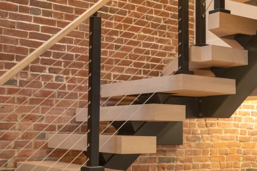 Light maple floating treads on a black mono stringer with cable railing against an exposed brick wall.