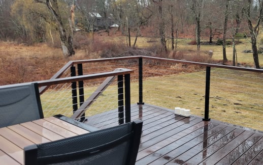 Black cable railing with Ipe top rail around a wet hardwood deck overlooking a wooded yard.
