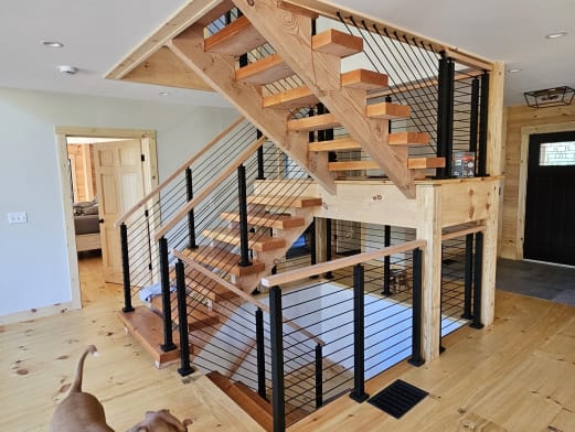 Open pine treads on wood stringers with black post cable railing around a switchback in a pine-floored cabin.