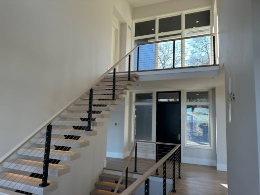 White oak floating treads with black-post cable railing and oak handrail climb to a bridge in a bright foyer.