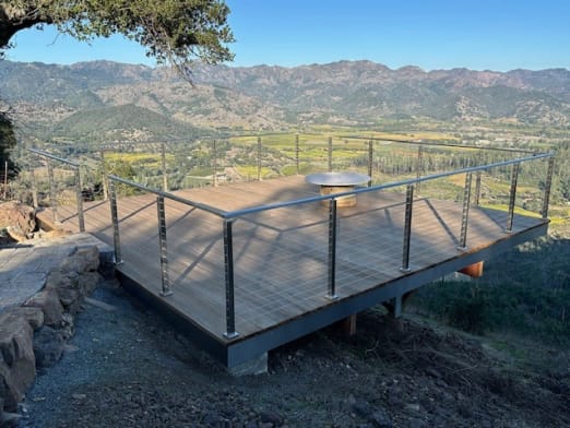 Brushed stainless cable railing around a wood deck perched above a vineyard valley under blue sky.