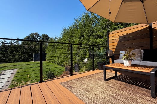 Black cable railing lines a warm-toned composite deck with outdoor seating, umbrella and privacy screen.