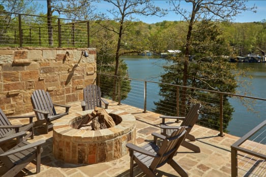 Burnished bronze cable railing lines a stone patio with a fire pit and Adirondack chairs above a lake.
