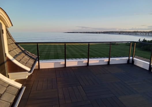 Black cable railing with a curved section borders a dark rooftop deck overlooking a lawn and the ocean at dusk.