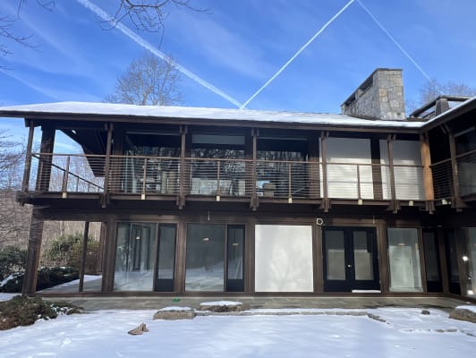 A dark-stained timber two-story home with a long upper balcony enclosed by horizontal stainless cable railing, snow on the ground.
