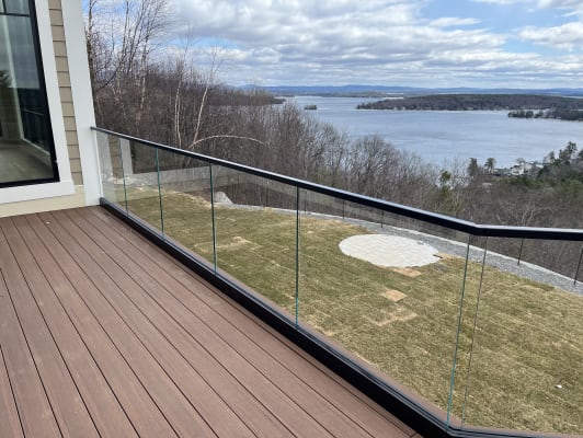 Base-rail frameless glass with a black cap edges a composite deck overlooking a wooded lake under clouds.