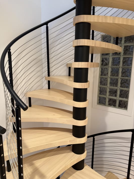 A maple spiral staircase with a curved black cable railing rising beside a glass block window.