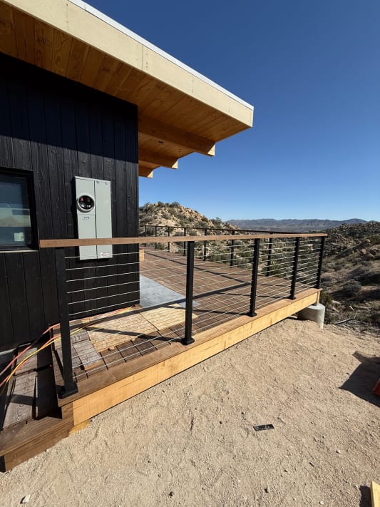 Black cable railing with a wood top rail wraps a timber deck on a dark-clad desert home.