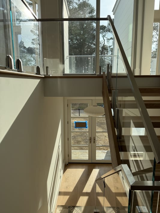 Frameless glass panels in stainless clamps with a square stainless handrail along an open oak stair facing tall windows.