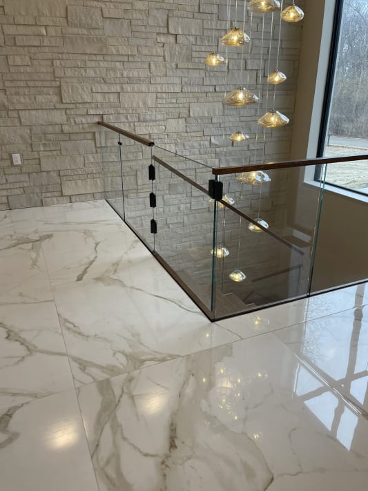 Frameless glass panels with a walnut handrail guard a stair opening on a glossy marble floor beneath a cascading glass pendant light.