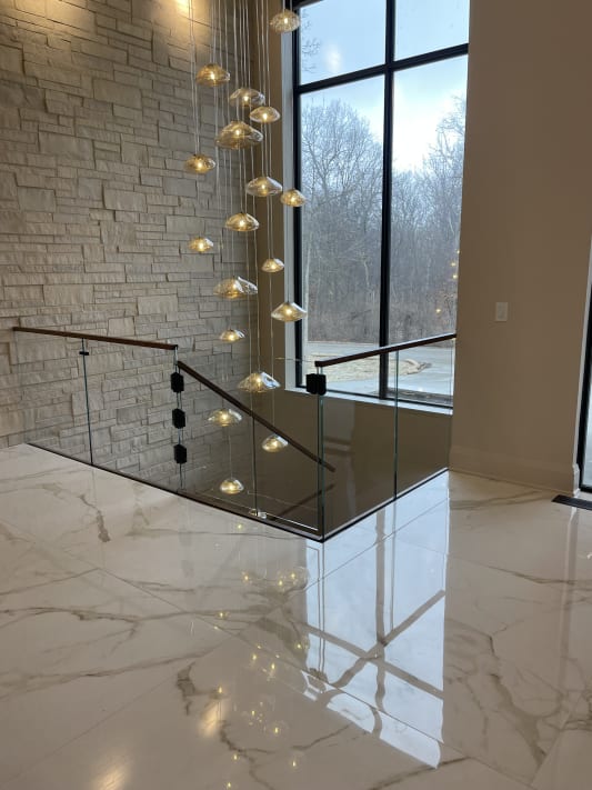 Frameless glass panels with a walnut handrail guard a stair opening on a glossy marble floor beneath a cascading glass pendant light.