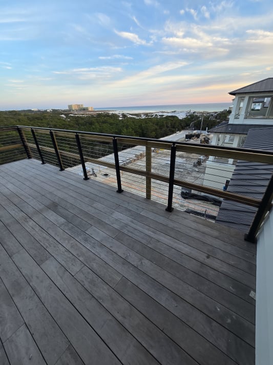 Black cable railing wraps a gray wood rooftop deck overlooking forest and a distant coastline at sunset.