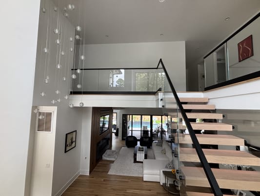 Floating white oak stair with glass railing rising to a glass-railed loft above a modern living room