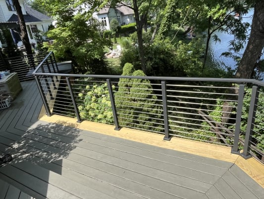 Black cable railing lines the edge of a gray deck overlooking a garden and lake.