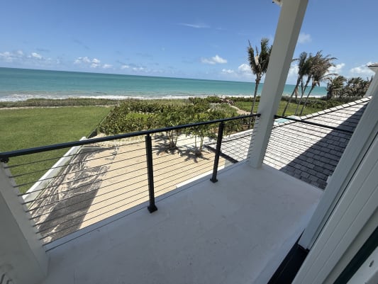 A covered balcony with black cable railing looks out over dunes, palms and the ocean.