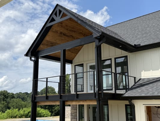 A black-post cable railing lines a covered balcony beneath a cedar-lined gable on a white board-and-batten home.