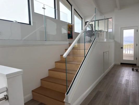 An oak stair rises beside frameless glass with a white handrail toward a glass-guarded loft and patio door.