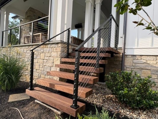 Chunky hardwood treads with a black cable railing rise to a covered porch above a stone wall.