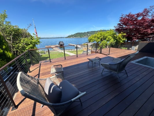 Woven lounge chairs sit on a hardwood deck edged by wood-capped cable railing with a lake beyond.