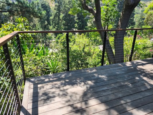 Wood-capped cable railing wraps a weathered hardwood deck surrounded by lush trees and greenery.