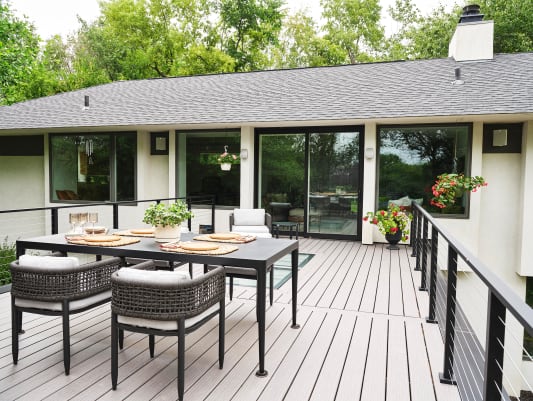 Black Express cable railing surrounds a pale composite deck with glass skylights and an outdoor dining set beside a white modern home.