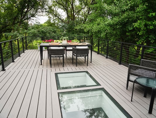 A composite deck with walk-on glass skylights and dining set wrapped in black cable railing among tall trees.