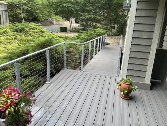 Legacy gray cable railing lines a composite walkway deck beside a shingled home and evergreen shrubs.