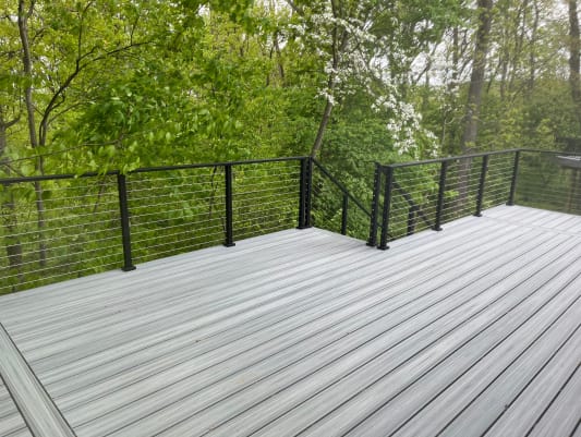 Black-post cable railing wrapping a gray composite deck overlooking dense green woods.