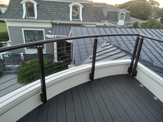 Curved black handrail and cable railing frame a dark-decked balcony above slate and metal rooftops.